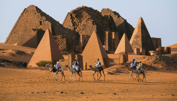 أشخاص يركبون عل الجِمال بجانب أهرامات النوبة في السودان