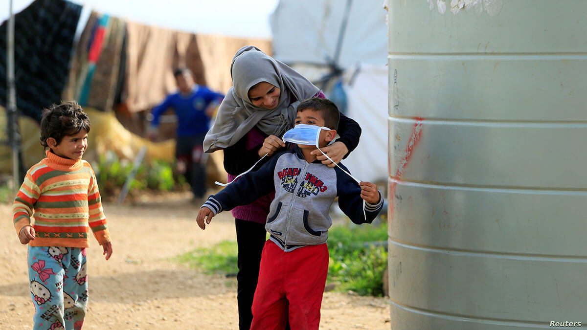 إمرأة تضع الكمامة على وجه أحد الأولاد في مخيم الوزاني للنازحين للوقاية من فيروس كورونا (رويترز)